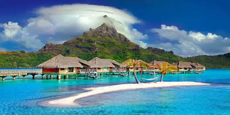 a beach with houses and a hammock on the water with Bora Bora in the background